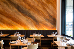 A modern restaurant interior with round and rectangular wooden tables set with white plates and wine glasses, dark upholstered booth seating, and a large warm-toned abstract wall mural behind them.