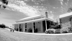 A black and white photograph of a single-story stone house with a wide covered porch, lawn in the foreground, and cloudy sky above.