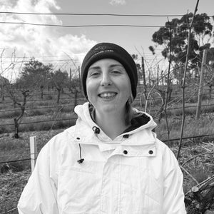 Shelby outdoors in a vineyard wearing a dark Two Hands beanie with white text and a white hooded jacket, photographed in black and white in our vineyard